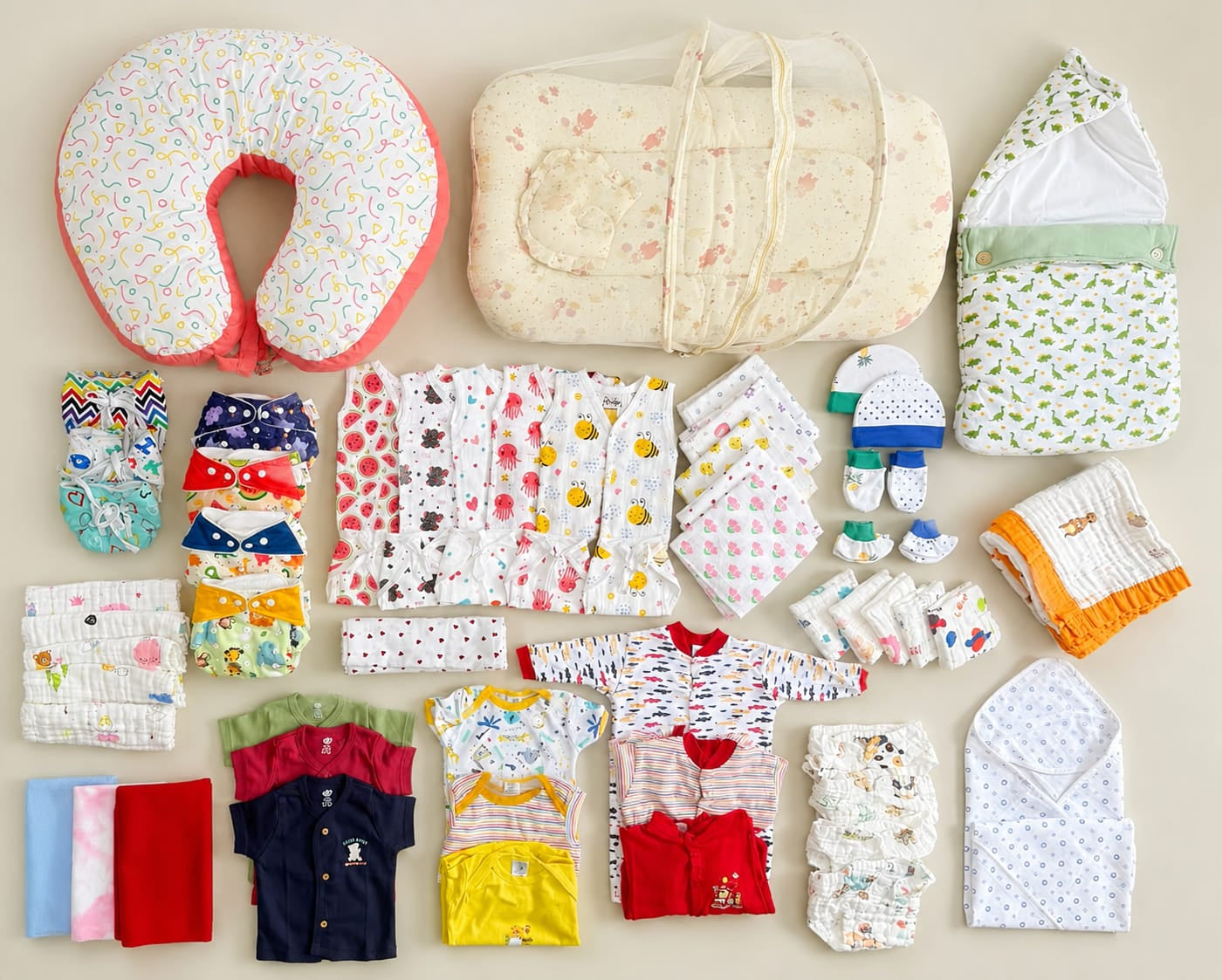 A flat lay of baby essentials and clothing items arranged neatly on a light beige background, including a patterned bassinet cover, a soft padded baby wrap, a U-shaped nursing pillow, folded baby clothes in multiple colors, and accessories.
