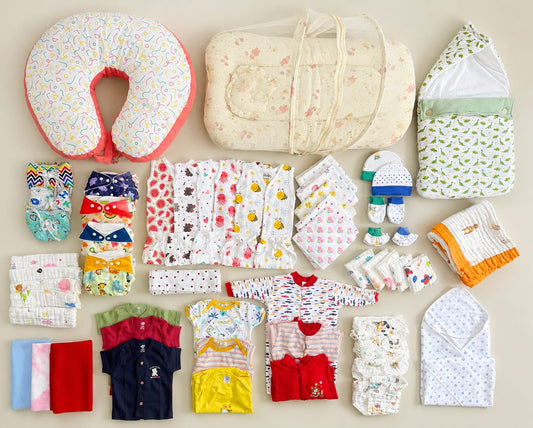 A flat lay of baby essentials and clothing items arranged neatly on a light beige background, including a patterned bassinet cover, a soft padded baby wrap, a U-shaped nursing pillow, folded baby clothes in multiple colors, and accessories.